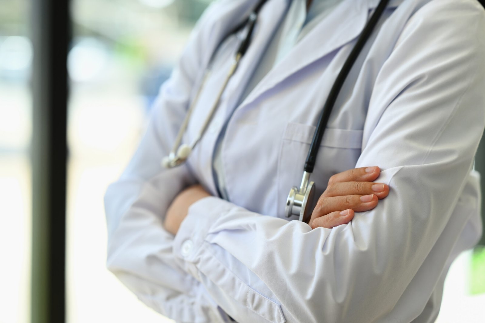 unrecognizable general practitioner standing with arms crossed standing near window in hospital